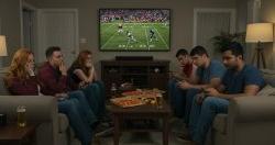 A group of people facing each other sitting on couches watching a football game. There is a table in the middle with pizza.